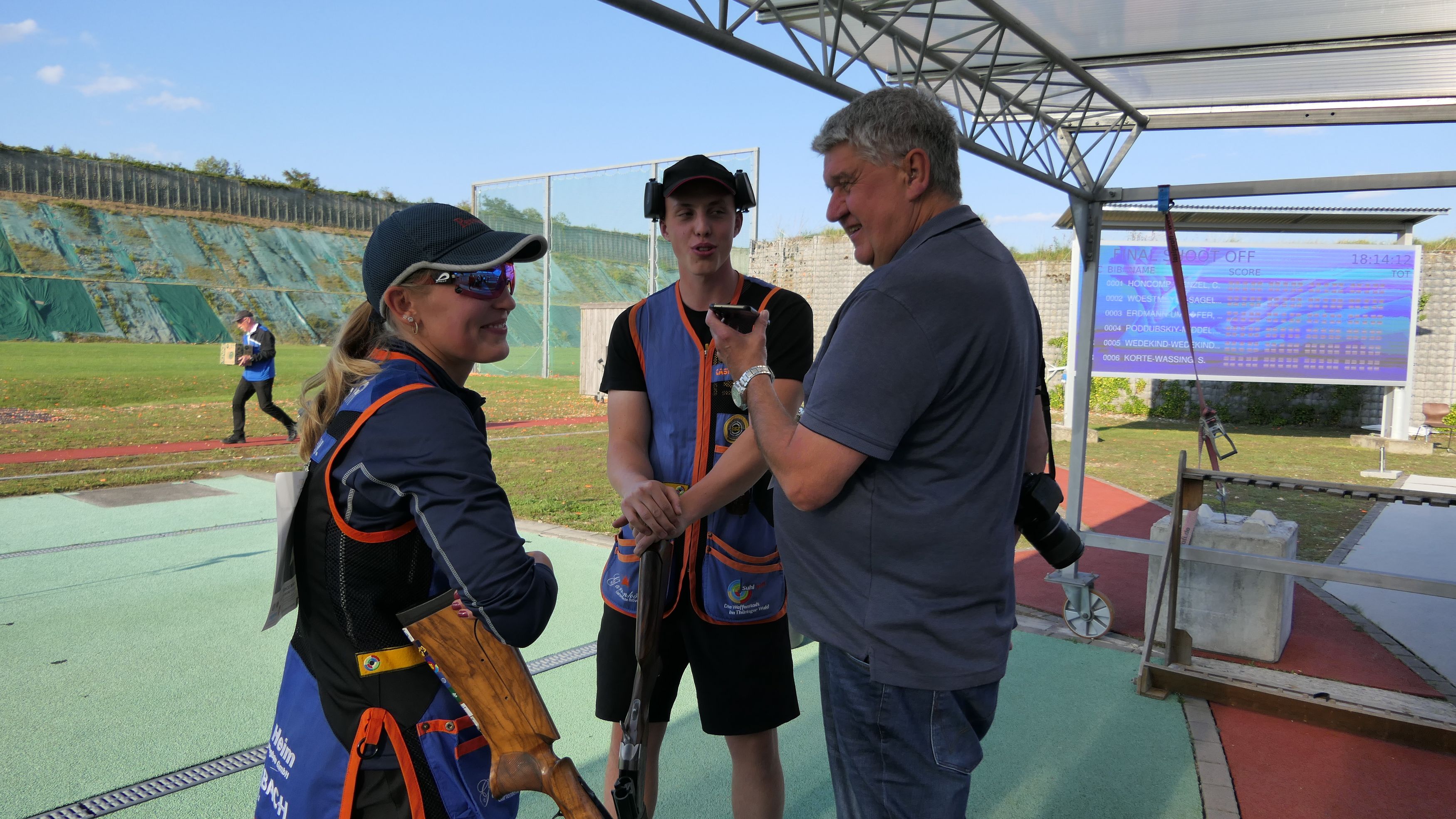 Foto: DSB / So kennt man Harald Strier: Fotokamera geschultert, Handy aufnahmebereit für Stimmenfang. Hier bei den Deutschen Meisterschaften mit dem Skeet-Duo Valentina Umhöfer und Magnus Erdmann.